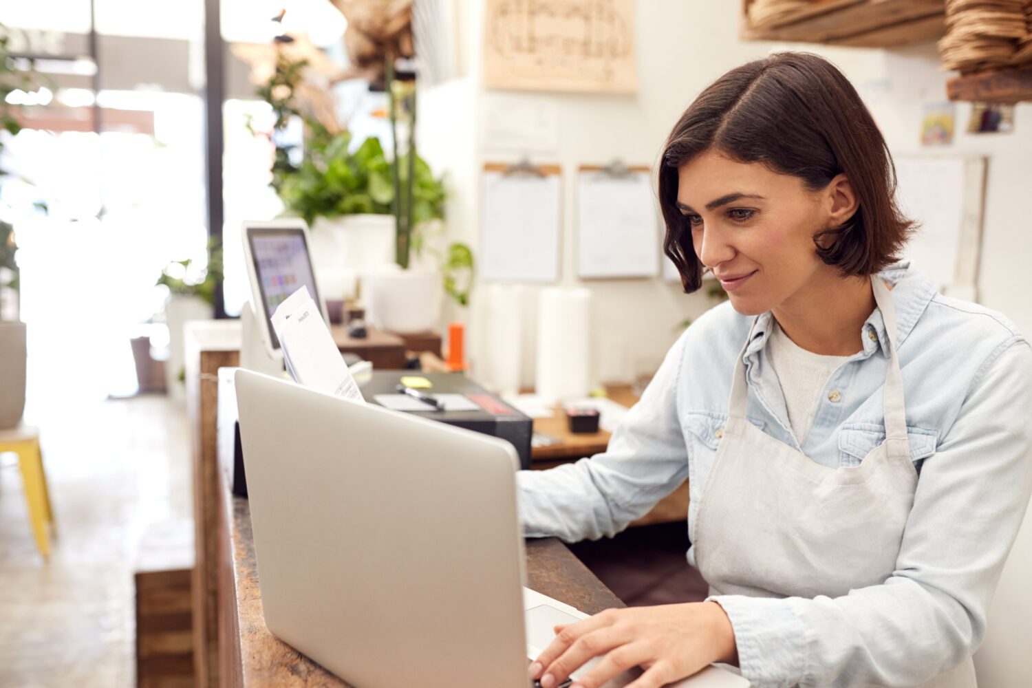 Female Sales Assistant Working On Laptop Behind Sales Desk Of Florists Store